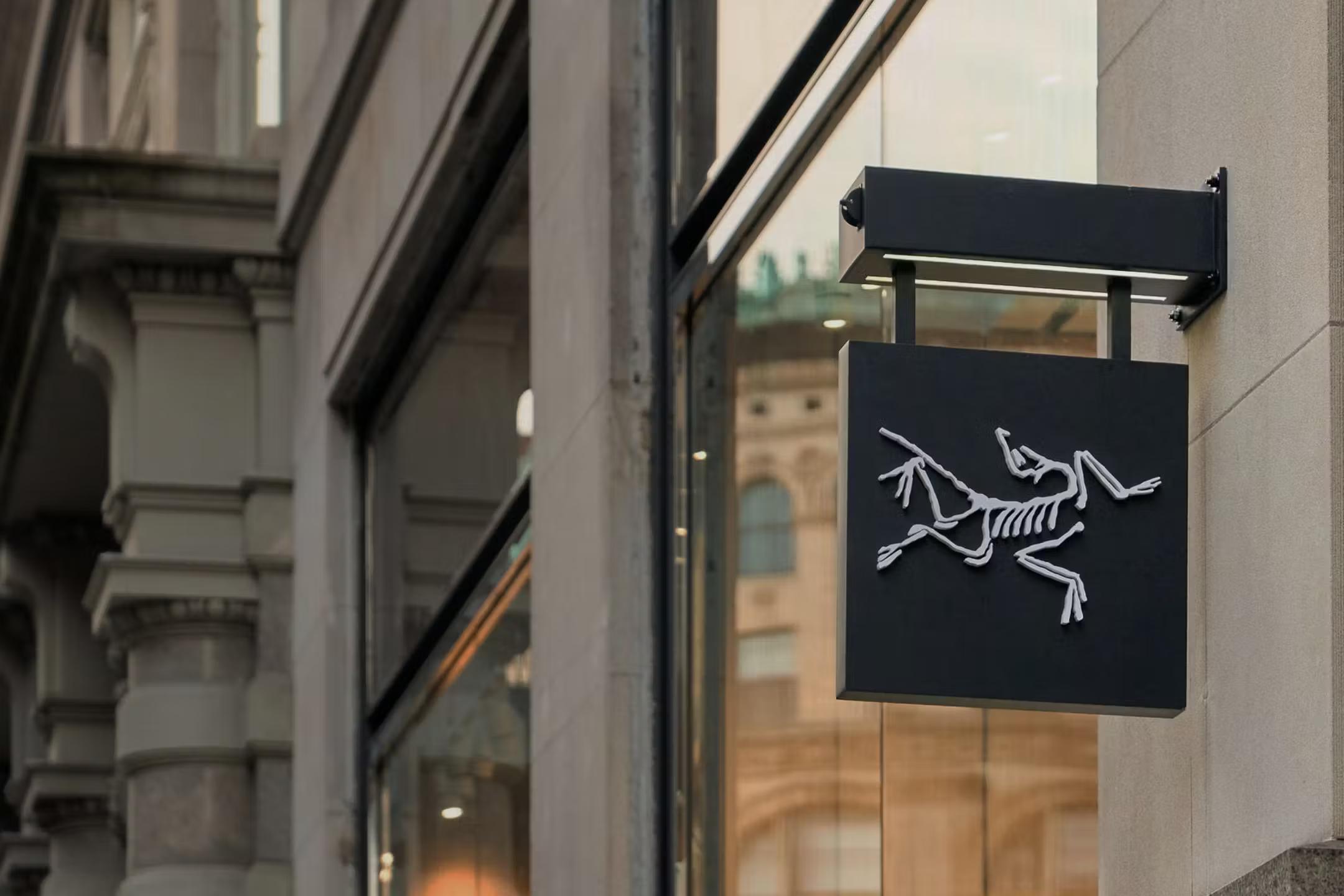 Black Arc'teryx sign with a white Archaeopteryx skeleton logo on a building facade.