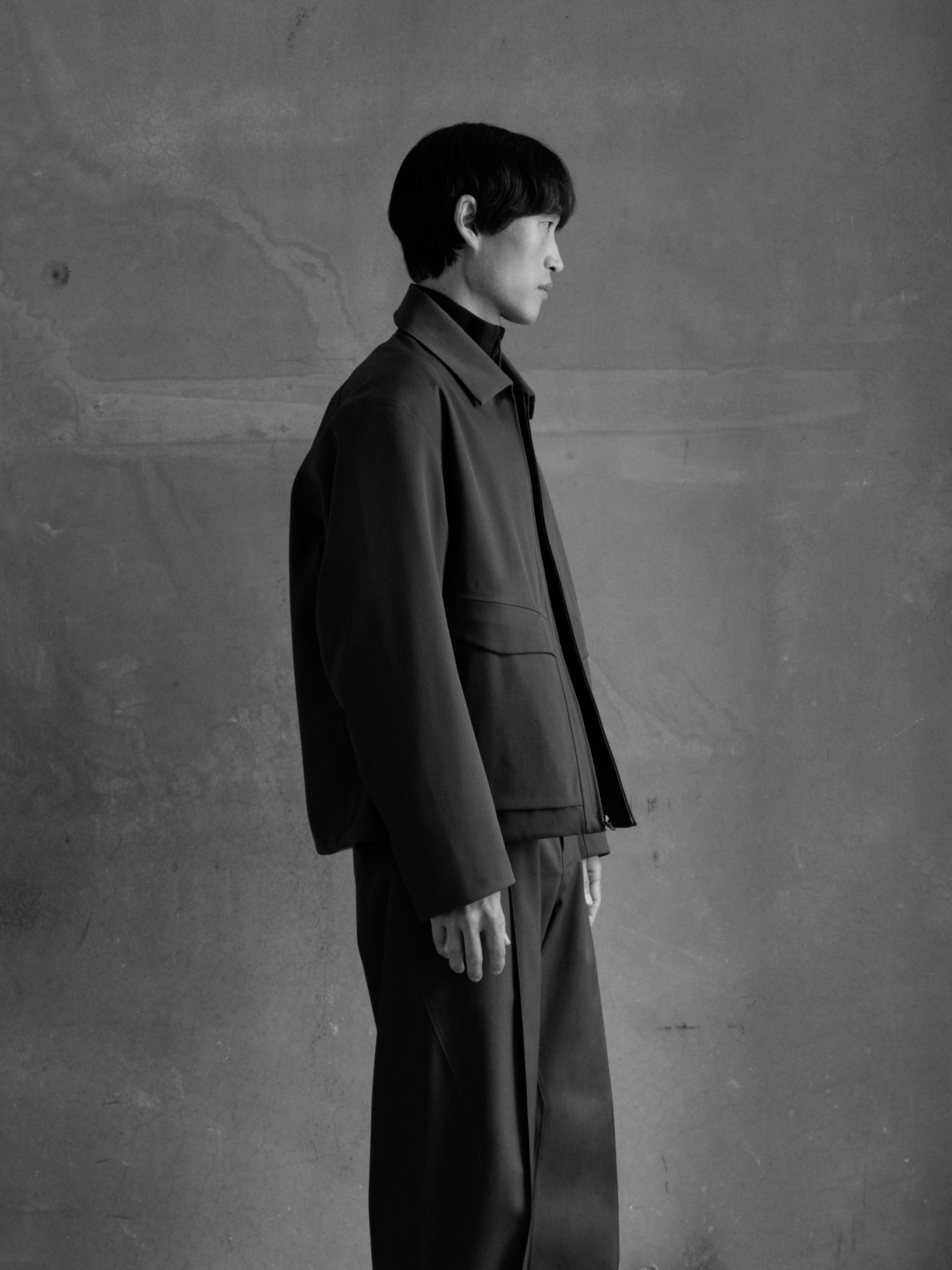 Monochrome profile shot of a man in a dark jacket, turtleneck, and wide-leg trousers against a textured wall.