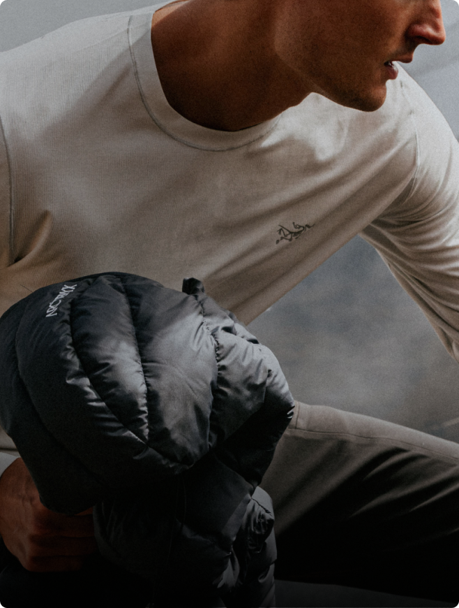 A man in a light long-sleeve Arc'teryx shirt holds a dark puffy Arc'teryx jacket.