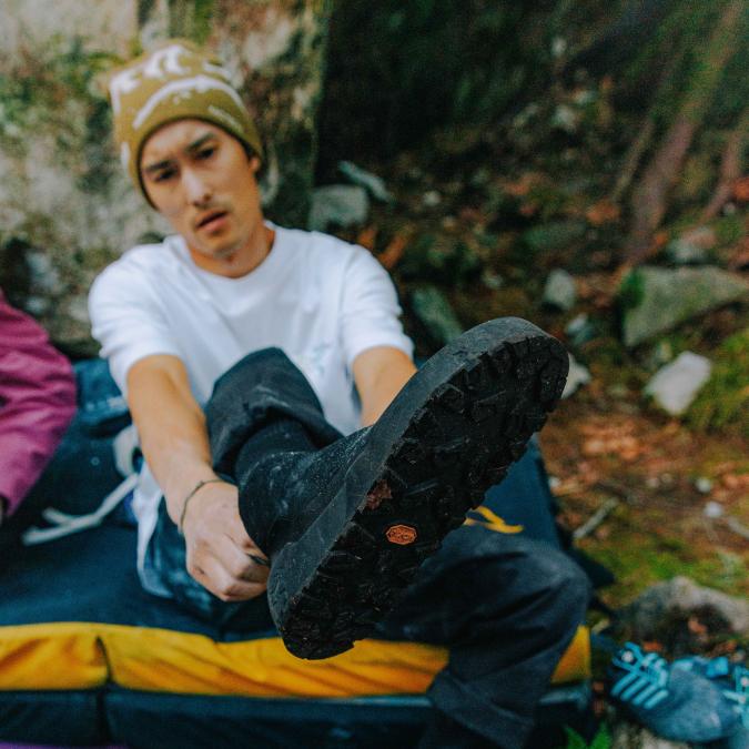 A person puts on a black boot, showing its Vibram sole, while sitting on a bouldering crash pad outdoors.