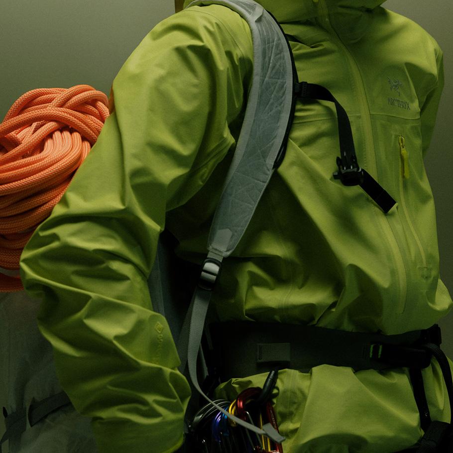 A person in a lime green jacket, wearing a backpack strap, with a coiled orange climbing rope and a harness with colorful carabiners.