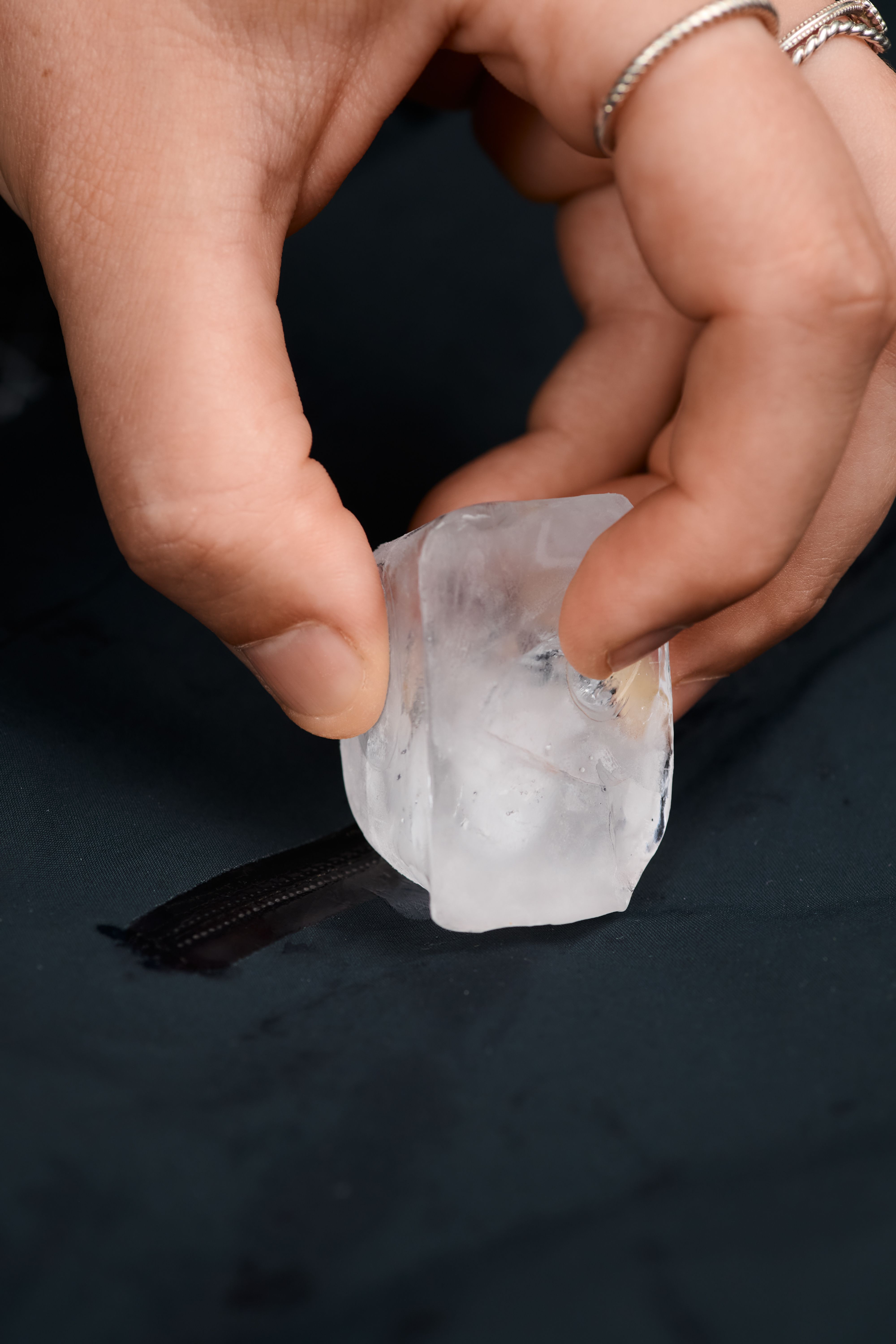 Hands hold a large ice cube, pressing it onto dark fabric with a zipper, causing a wet streak.