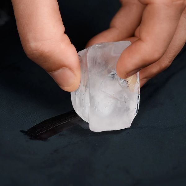 Hands hold a large ice cube, pressing it onto dark fabric with a zipper, causing a wet streak.