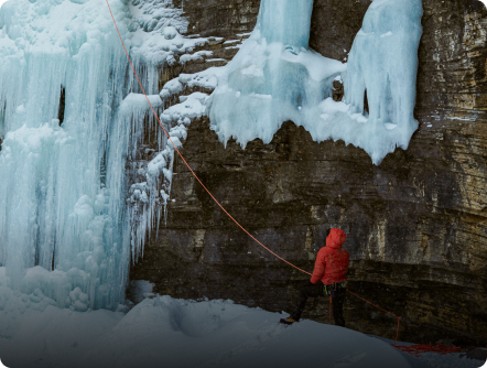 An ice climber in a red jacket stands on snowy ground at the base of a large frozen waterfall.