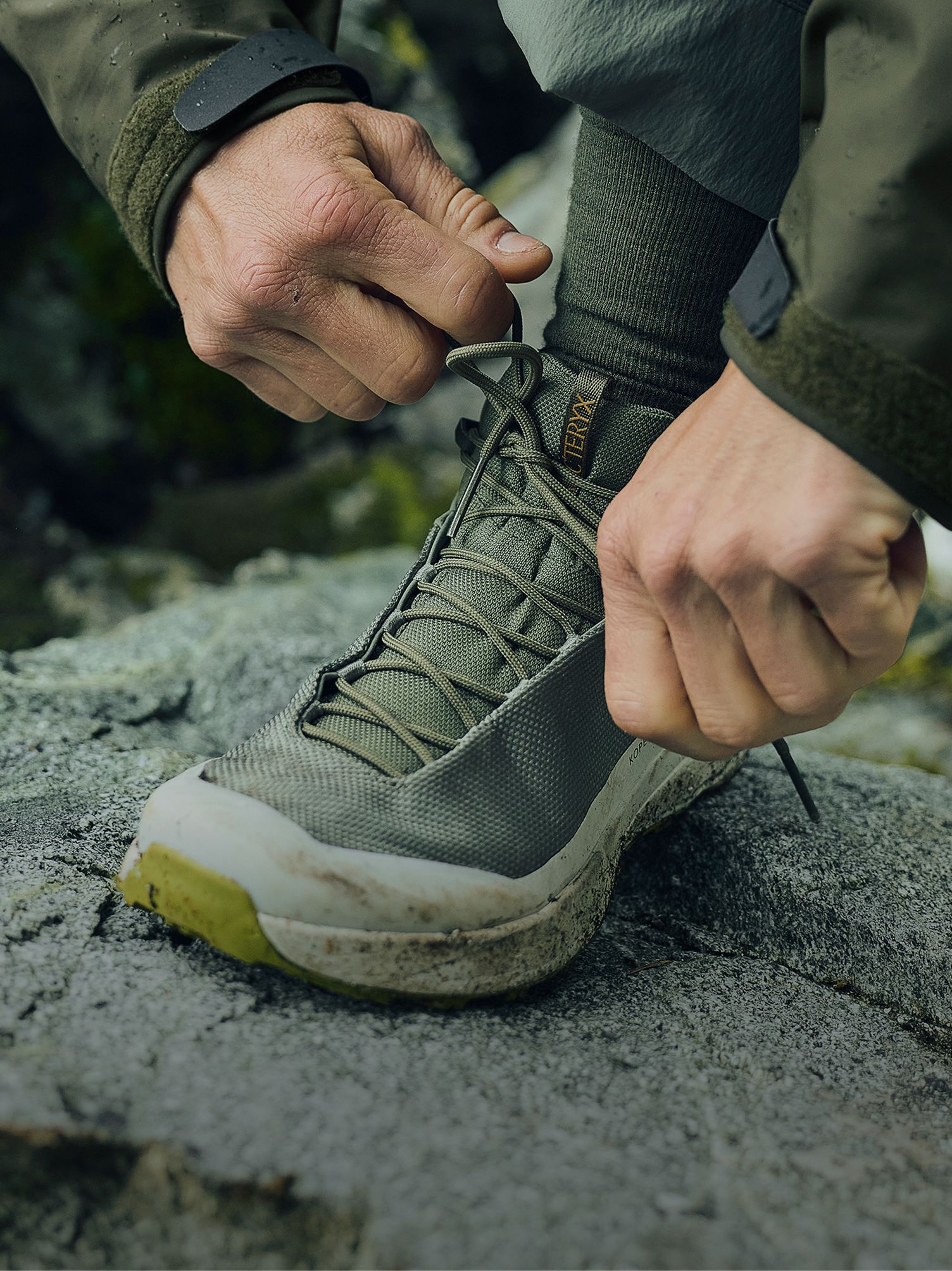 A person's hands tie the laces of a dirty olive green Arc'teryx hiking shoe on a rocky surface.