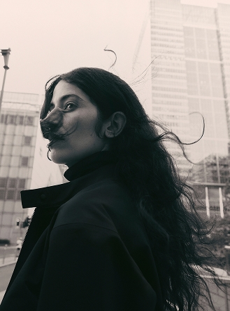 Monochrome profile of a woman with long, wind-blown hair looking up, against a blurred city background.