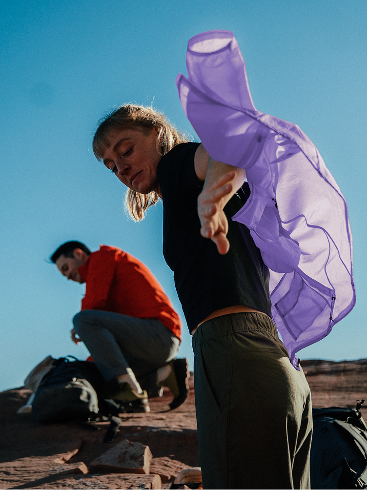 A woman adjusts a purple jacket, viewed from below against a blue sky, while a man crouches behind her.