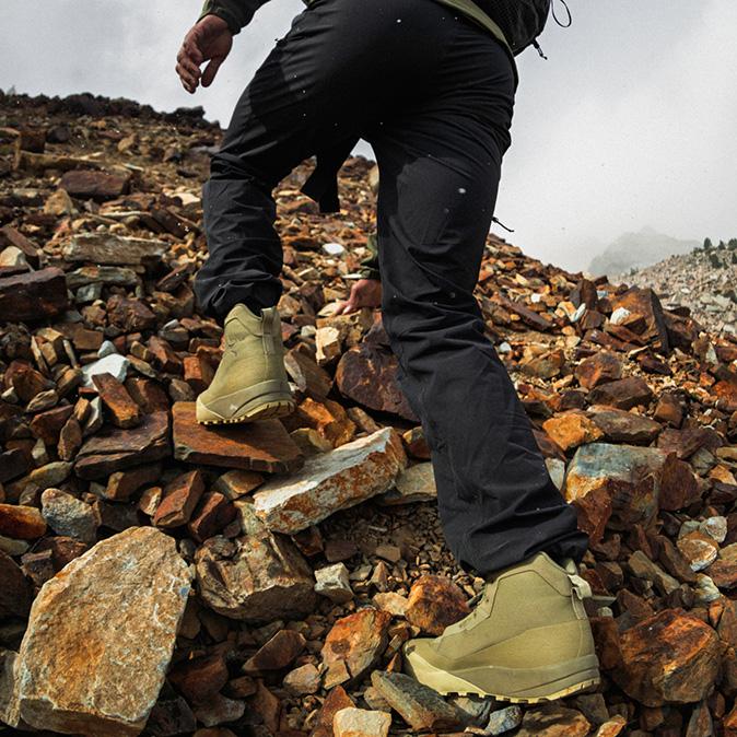 A person in black pants and tan hiking boots walks uphill on a steep, rocky trail.