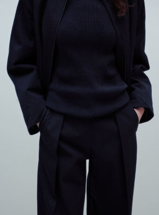Person in a navy ribbed top, open jacket, and trousers with front panels, hands in pockets.