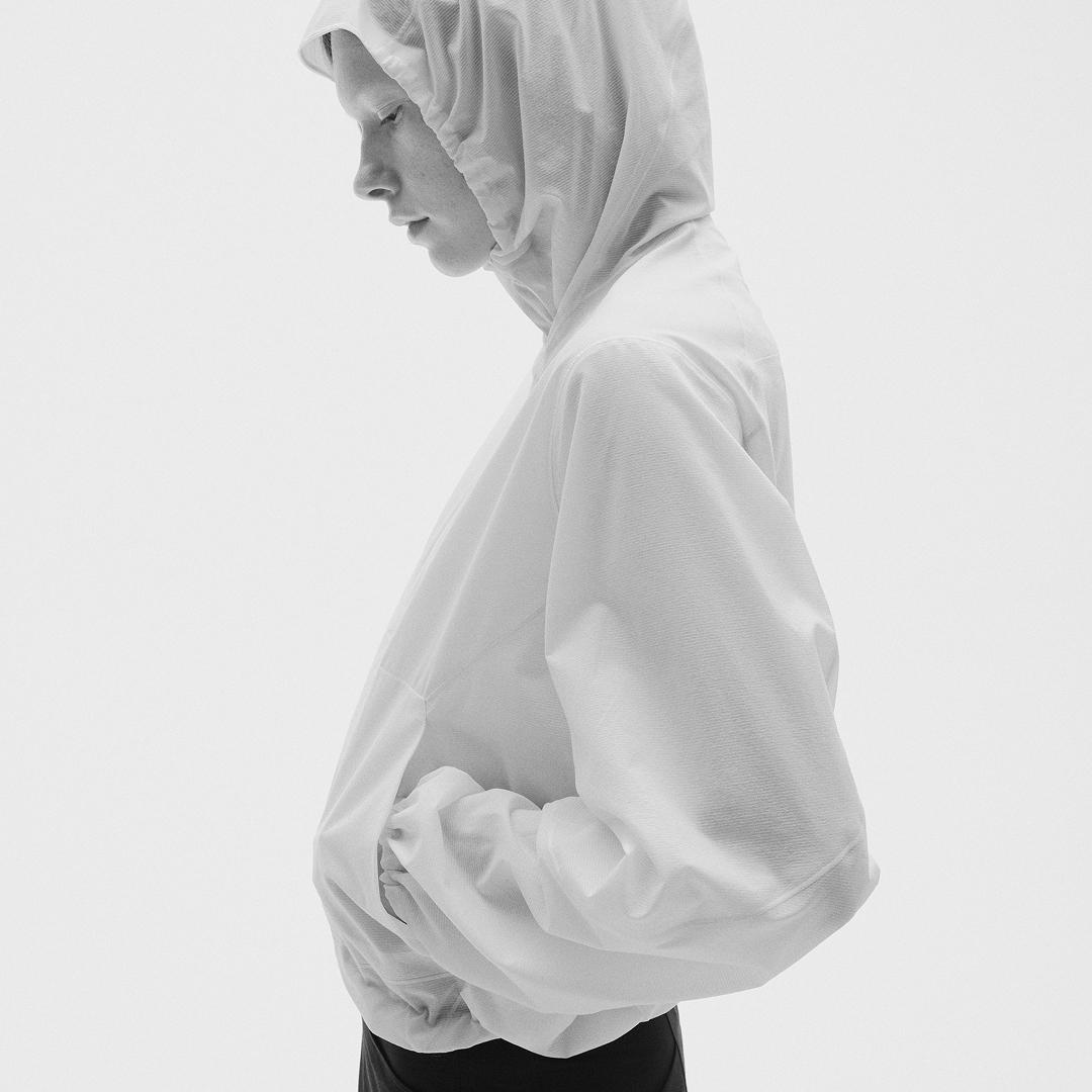 A person in a light hooded jacket, looking down with hands in pockets, in black and white.