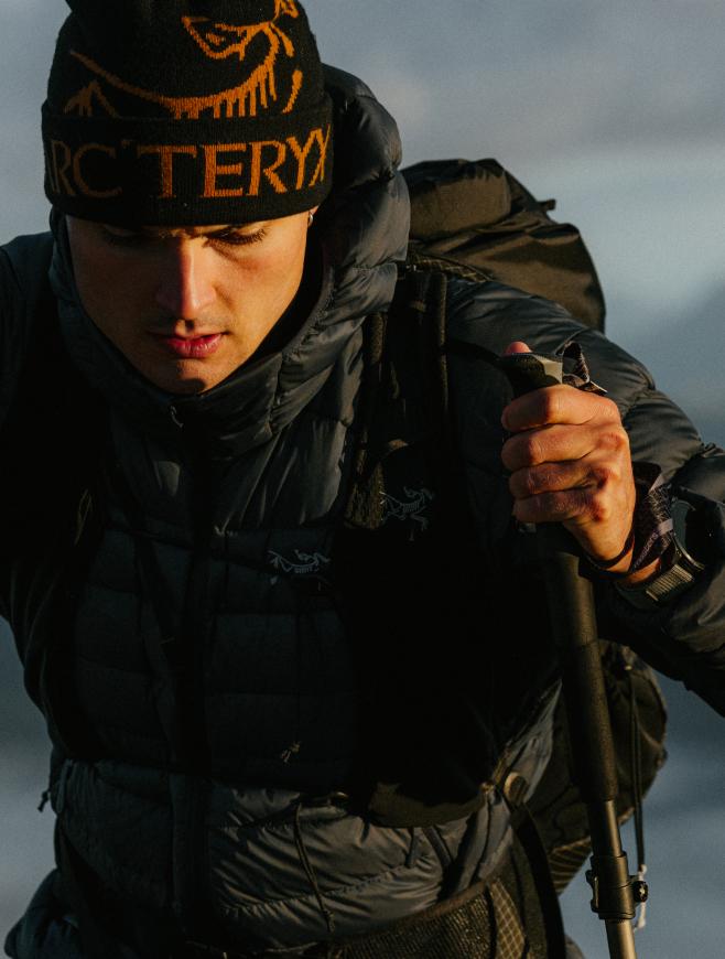 A man in a black Arc'teryx beanie and dark puffy jacket, carrying a backpack and holding a trekking pole while hiking.