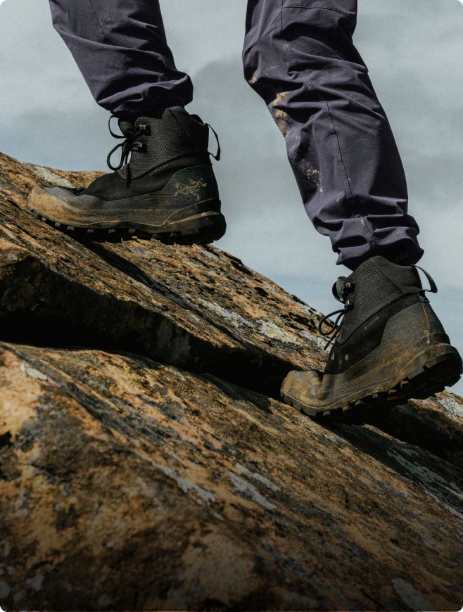 Dirty black hiking boots and dark pants climbing a rocky slope.
