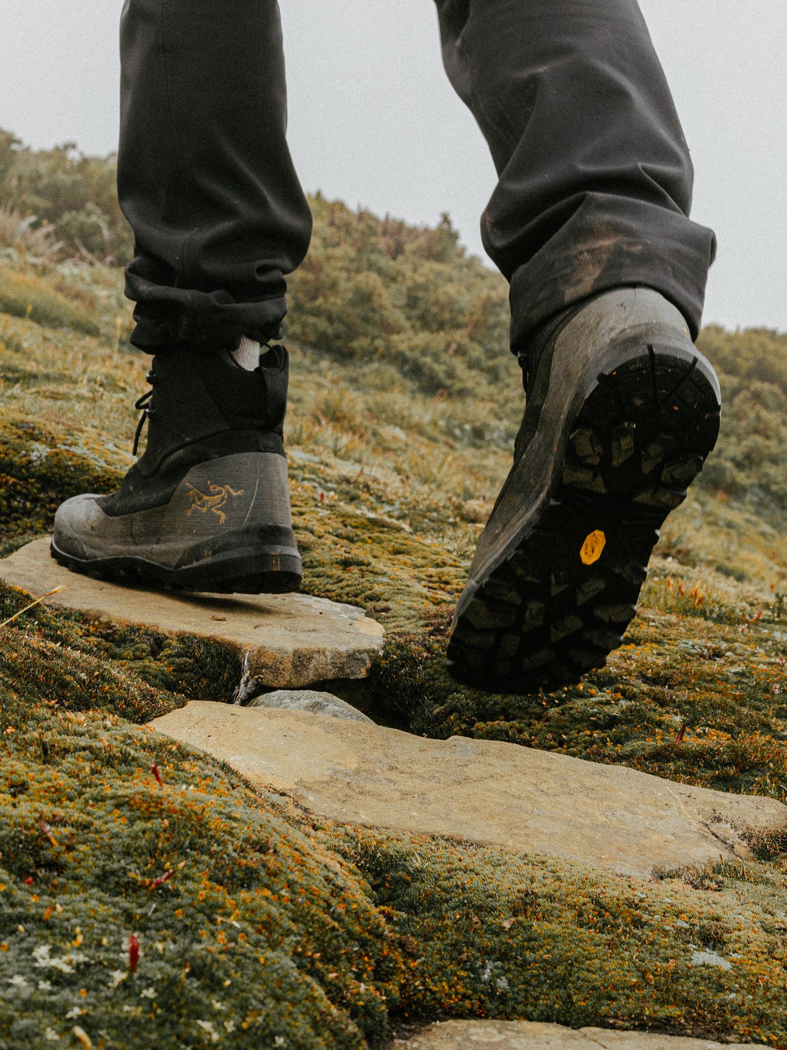 A person's feet in Arc'teryx hiking boots step on a stone path through moss, showing a Vibram sole.