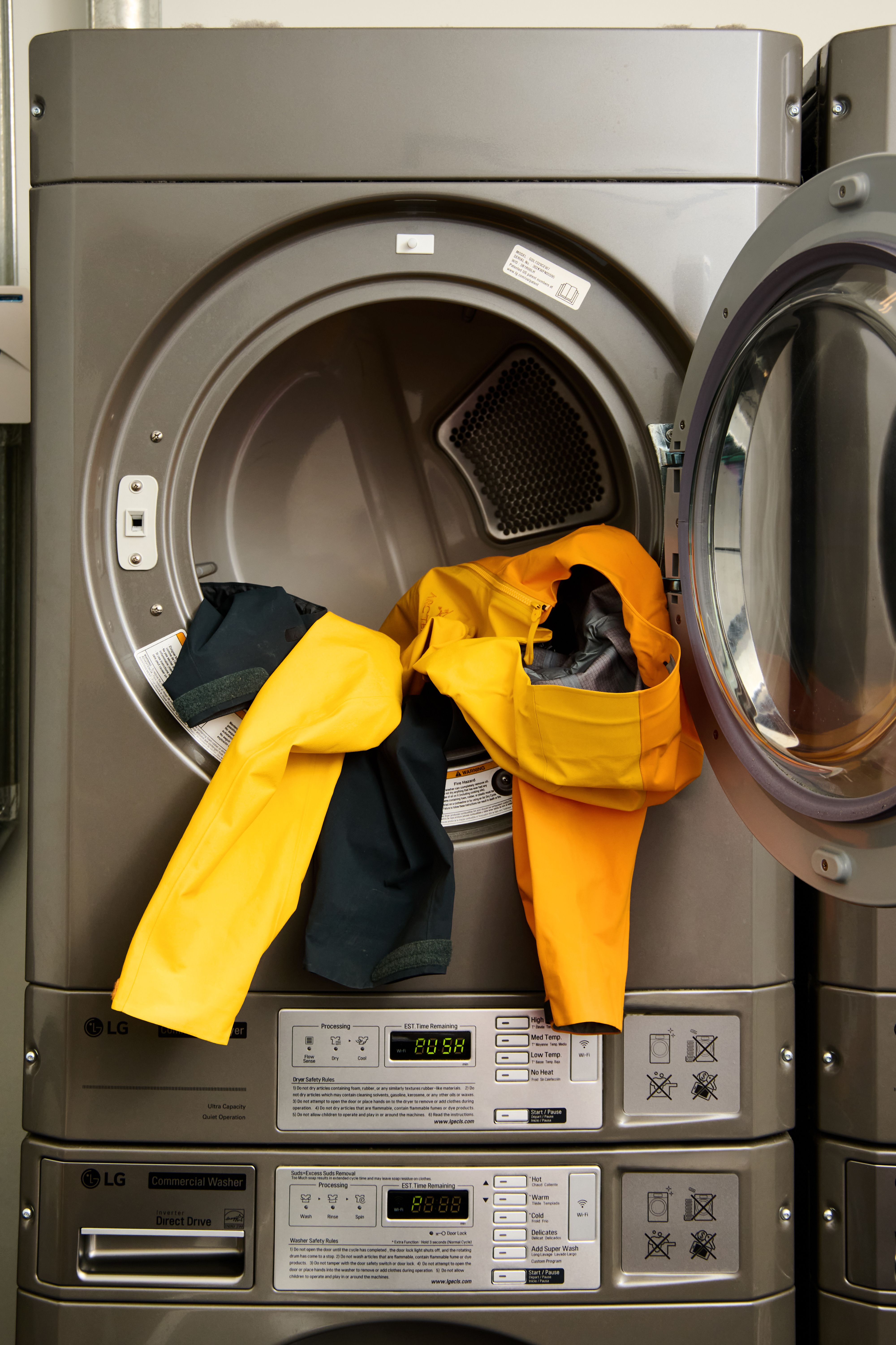 An open commercial dryer with yellow and dark clothes spilling out, stacked on top of a commercial washer.