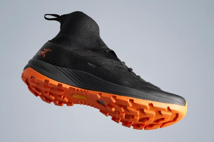 A black Arc'teryx trail shoe with a bright orange lugged sole and a high sock-like ankle, floating against a light background.