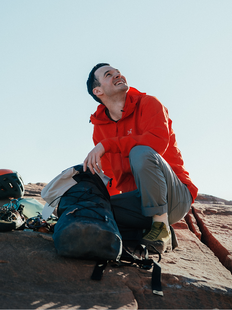 A smiling man in a red jacket crouches on rocky ground, looking up, with a backpack and climbing gear next to him.