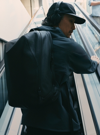 Man with black backpack, cap, and headphones riding an escalator.