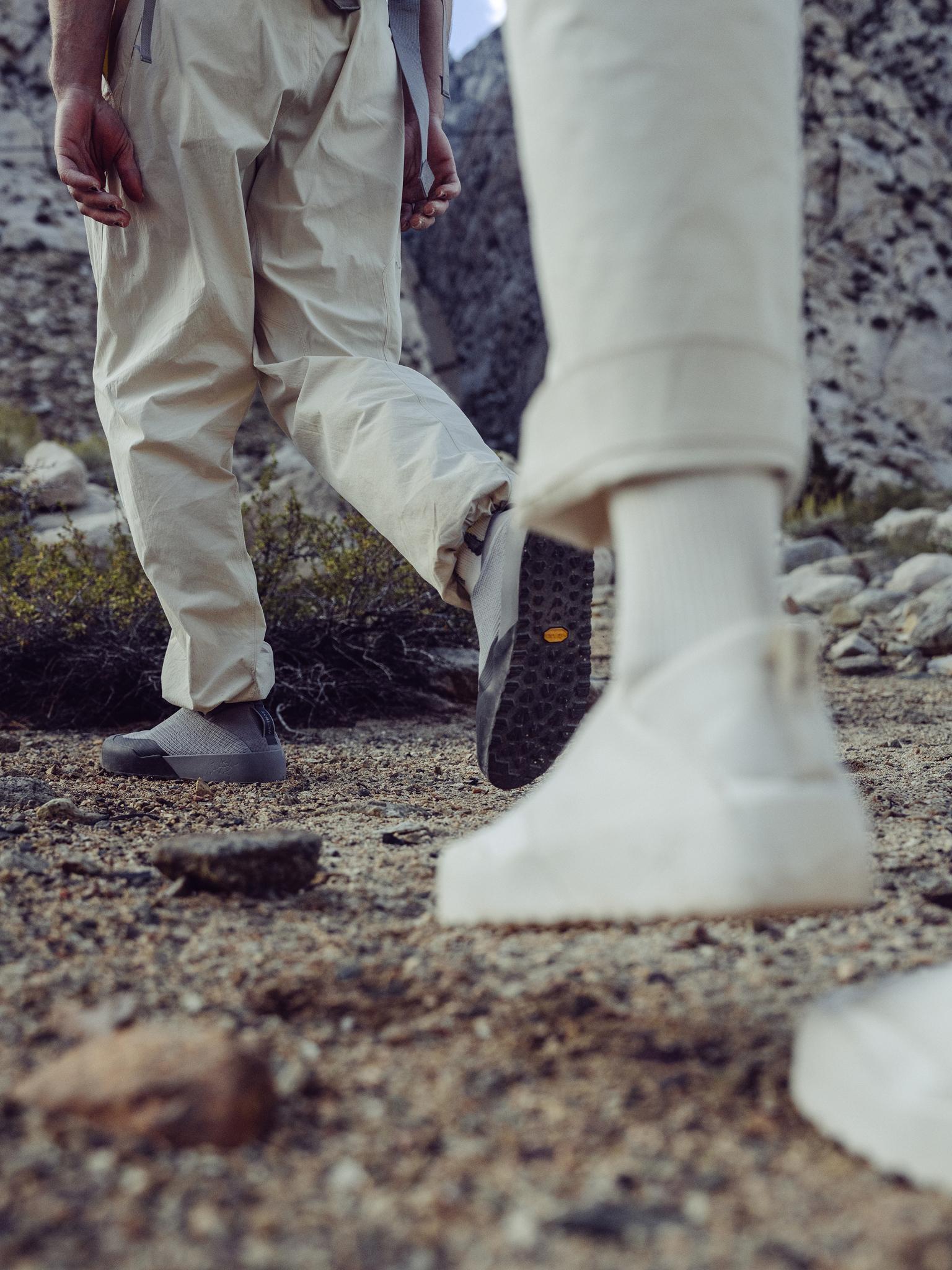 Two people wearing light-colored pants and outdoor shoes walk on a gravel path, one shoe revealing a black Vibram sole.