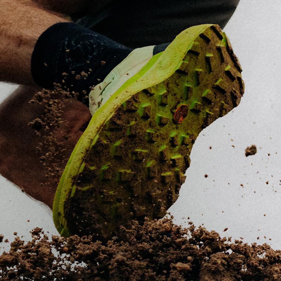 A dirty green trail running shoe sole kicks up soil.