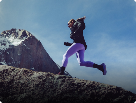 Woman in purple leggings leaping over rocky terrain with a snow-capped mountain in the background.