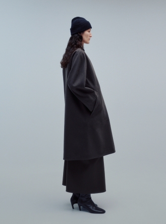 Woman in profile wearing a dark beanie, a dark grey loose coat, a matching midi skirt, and black boots.