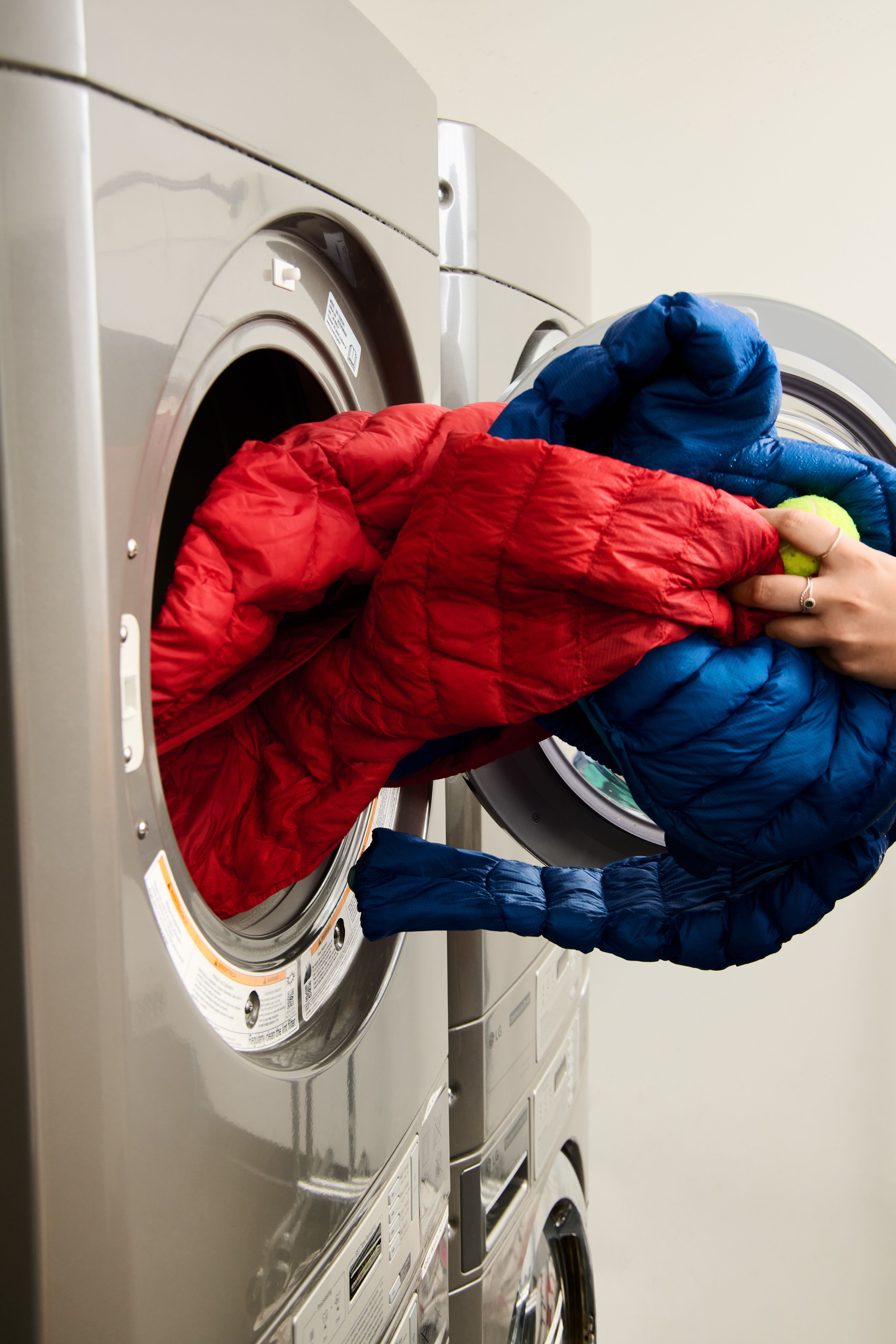 Hand inserting red and blue puffer jackets and a tennis ball into a front-load washer.