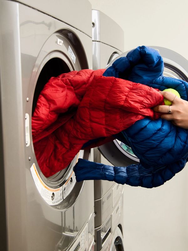Hand inserting red and blue puffer jackets and a tennis ball into a front-load washer.