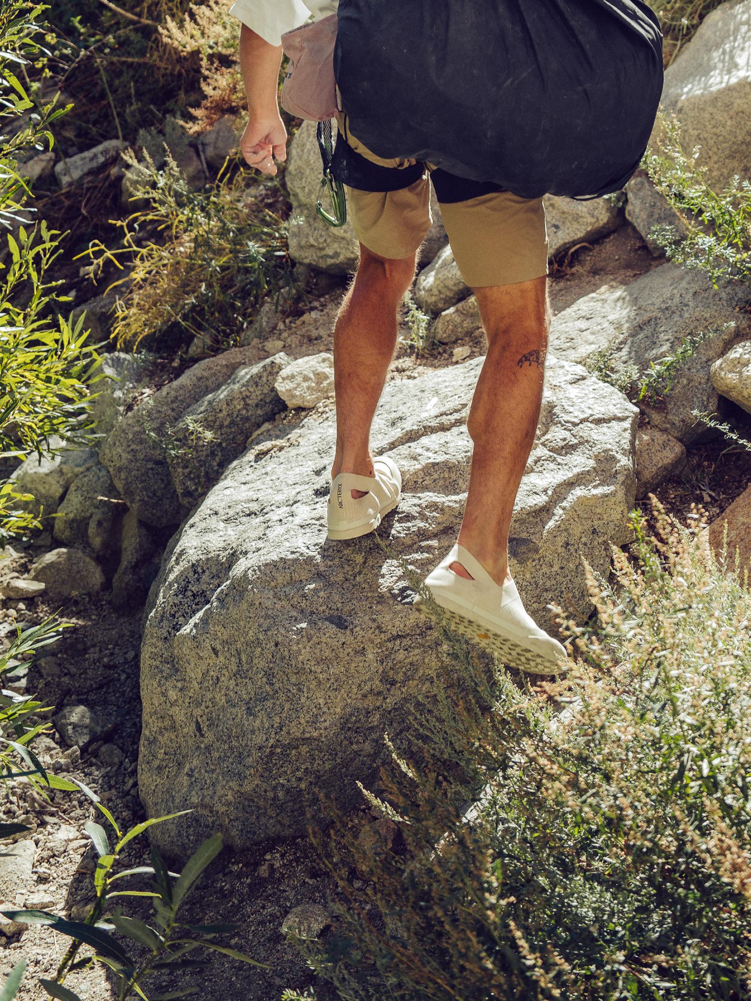 A person climbs a rocky trail, seen from behind, wearing khaki shorts, a harness, and light-colored shoes.