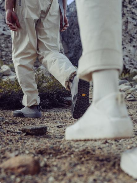 Two people in light-colored pants and minimalist shoes walk on a rocky path, one grey shoe showing a Vibram sole.