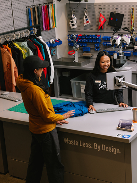 An employee at a repair station assists a customer with a blue jacket, with 'Waste Less. By Design.' visible on the counter.