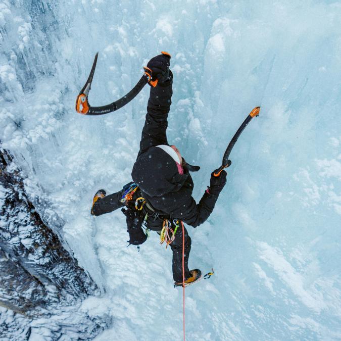Person ice climbing a vertical ice wall with two ice axes and crampons.