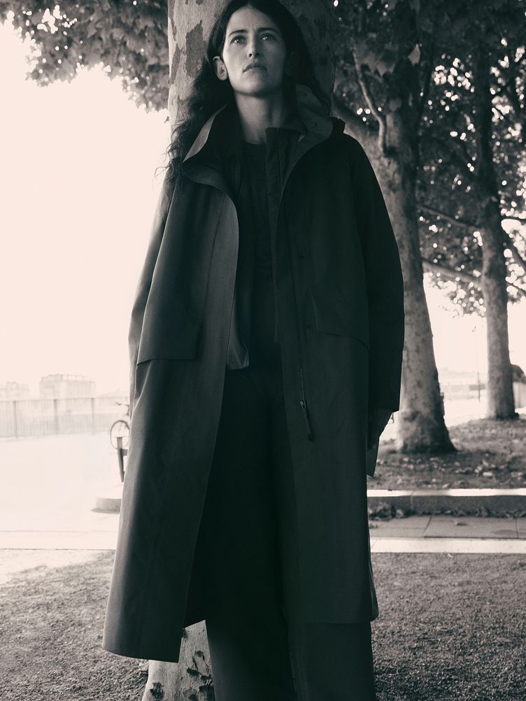 Black and white photo of a woman in a long coat standing by a tree, looking up.