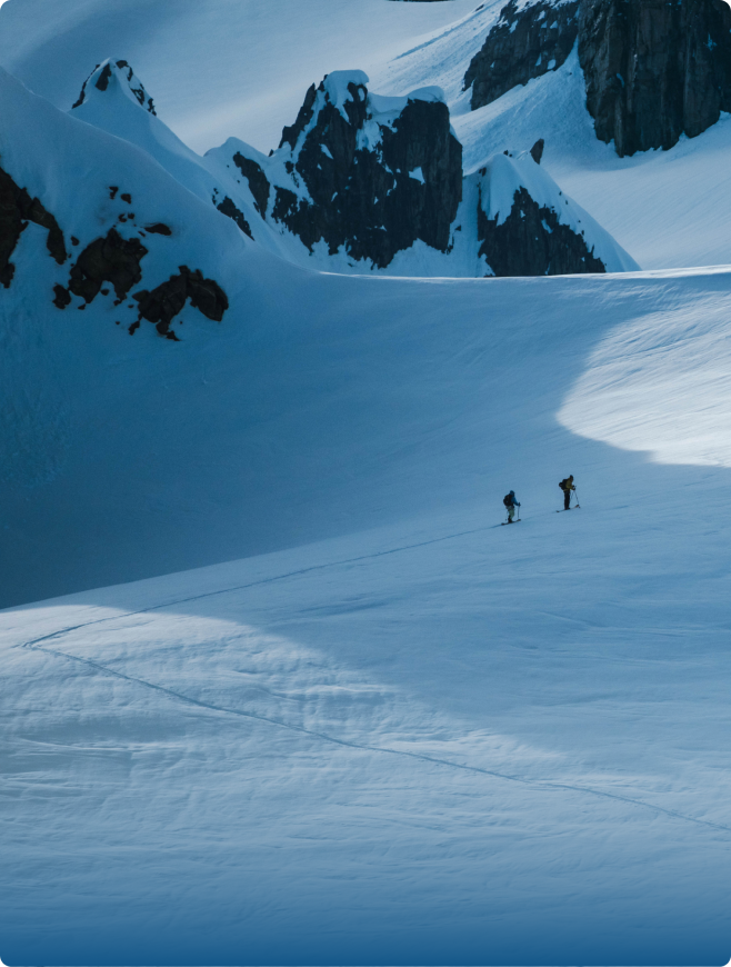 Two ski tourers traverse a vast, snow-covered mountainscape with jagged peaks.