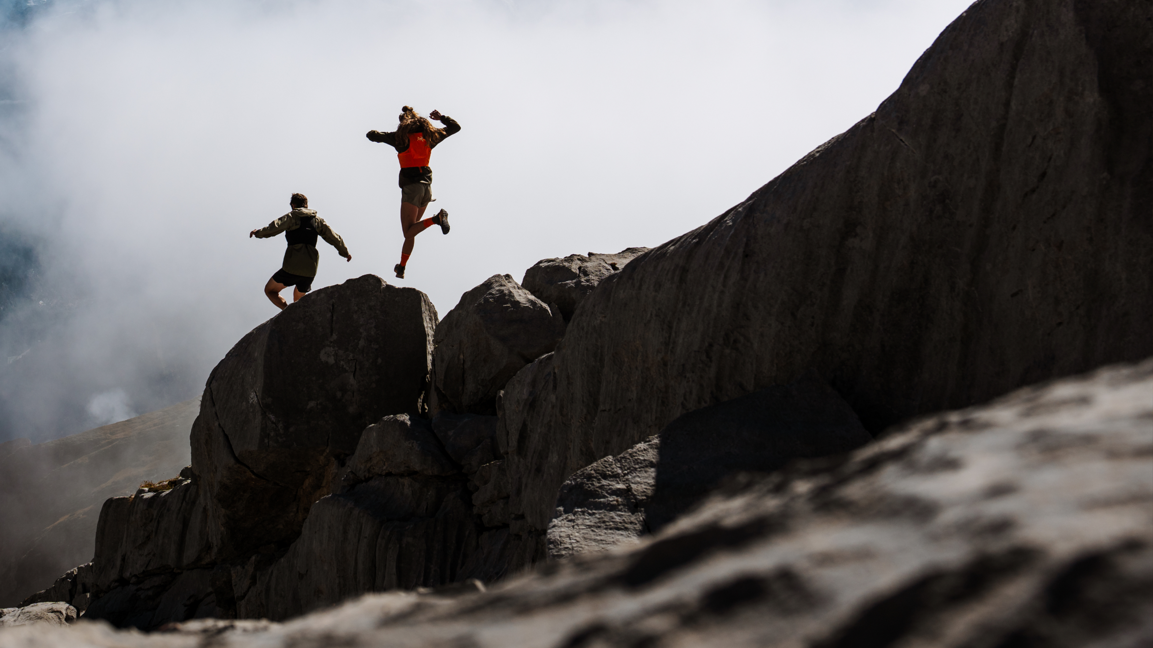Two people on rocky terrain, one leaping between boulders under a cloudy sky.
