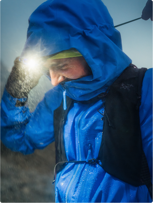 A person in a blue rain jacket with a bright headlamp, wet from water droplets.