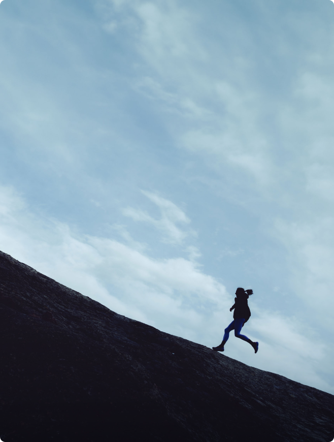 A silhouetted person runs up a steep dark slope under a cloudy sky.