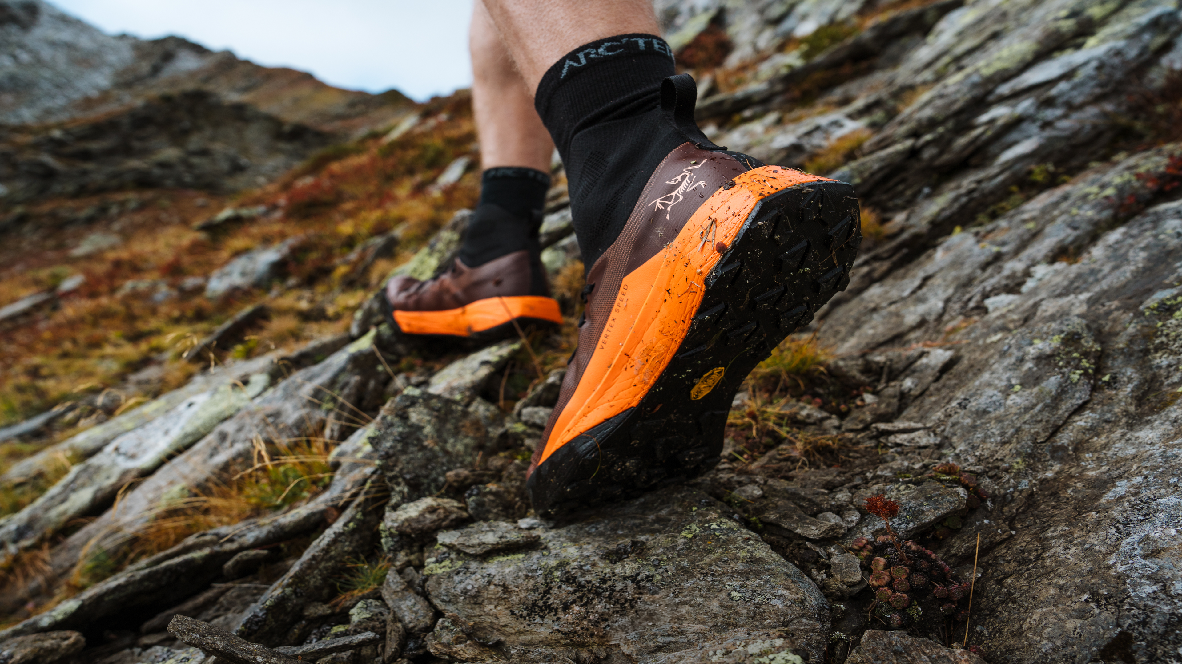 A person hikes uphill on a rocky trail wearing brown and orange trail running shoes.