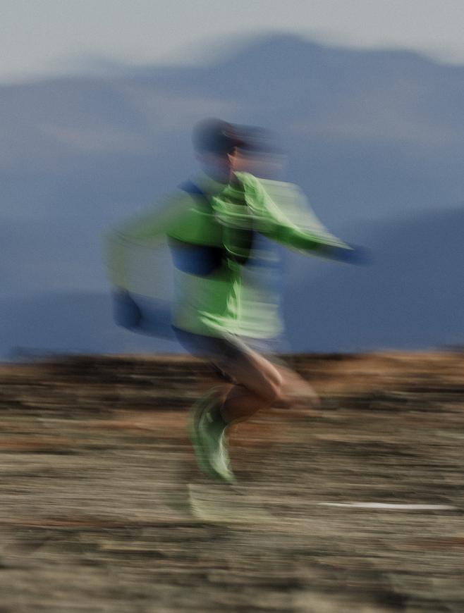 A motion-blurred runner in a bright green top races across a rocky trail with mountains in the distance.