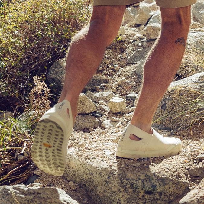 A person wearing light-colored trail shoes walks on a rocky path.