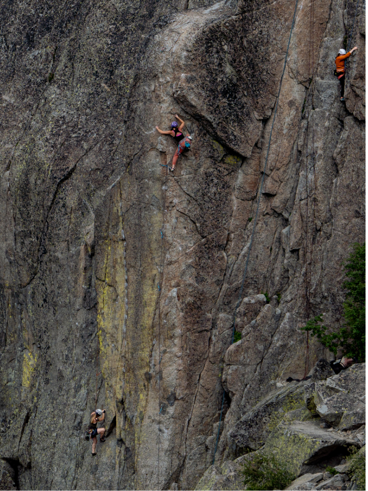 Three climbers ascend a tall, textured rock face with patches of lichen.