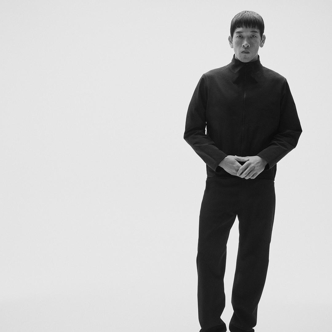 Monochrome three-quarter shot of a man in a black collared jacket and pants, hands clasped, looking directly forward.