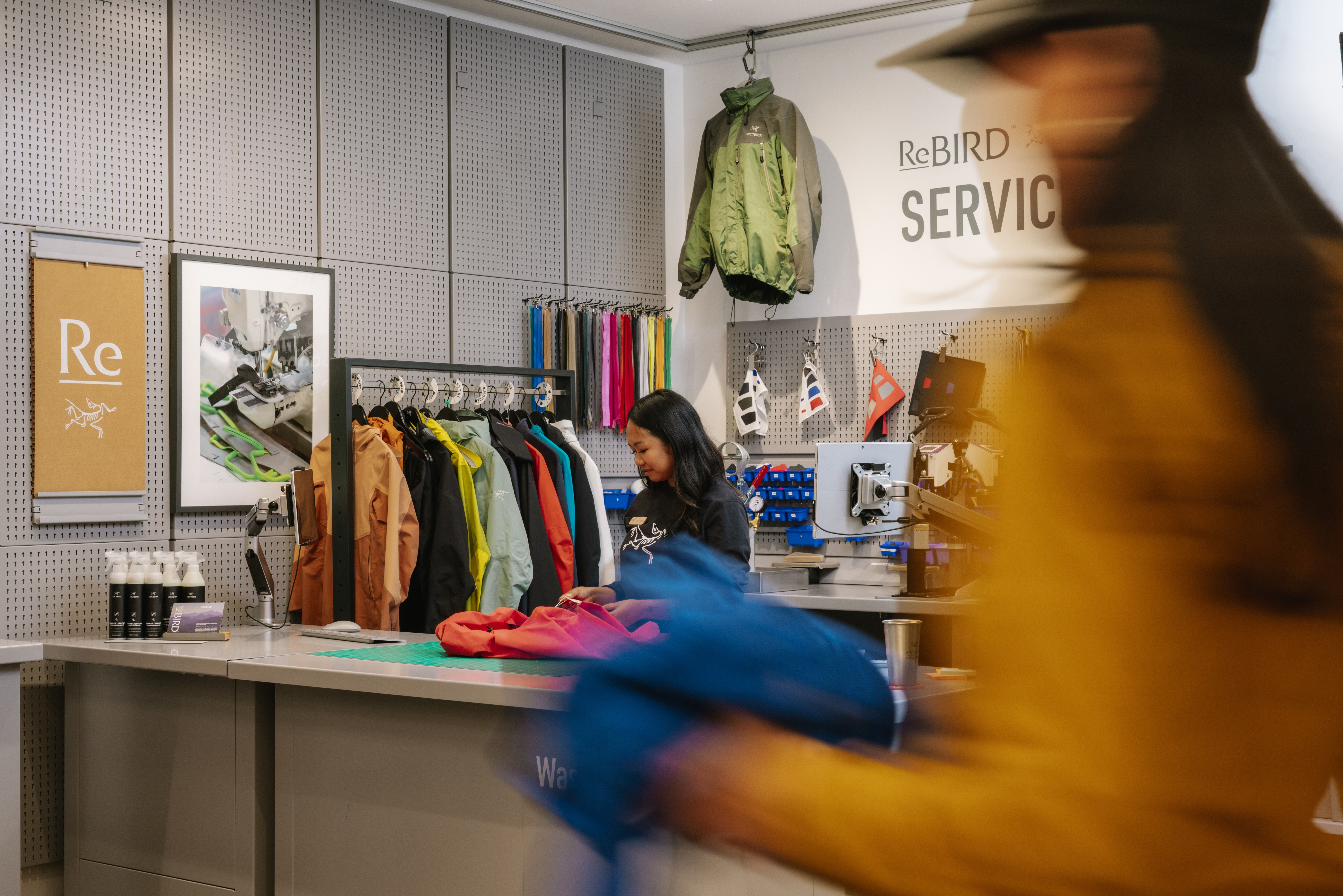 An employee at an Arc'teryx ReBird service counter mending clothing, with colorful jackets on display and a blurred customer walking by.