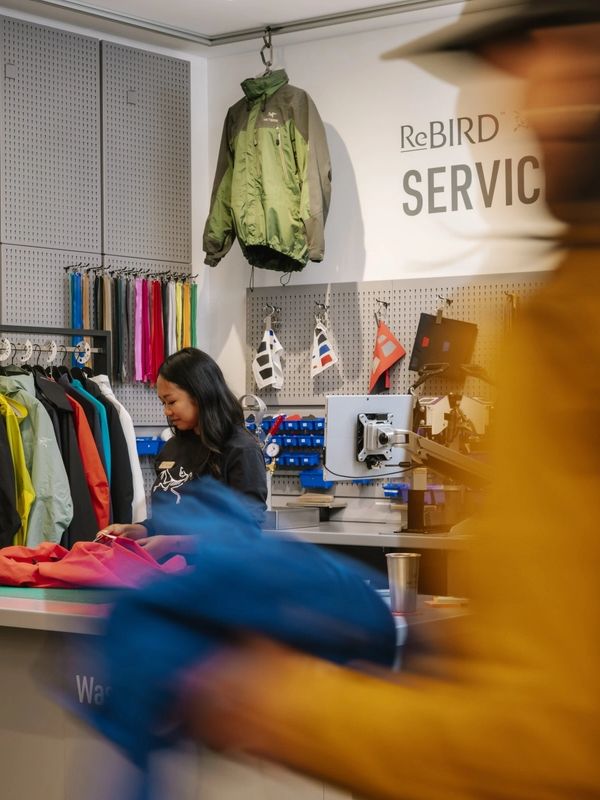 An employee at an Arc'teryx ReBird service counter mending clothing, with colorful jackets on display and a blurred customer walking by.