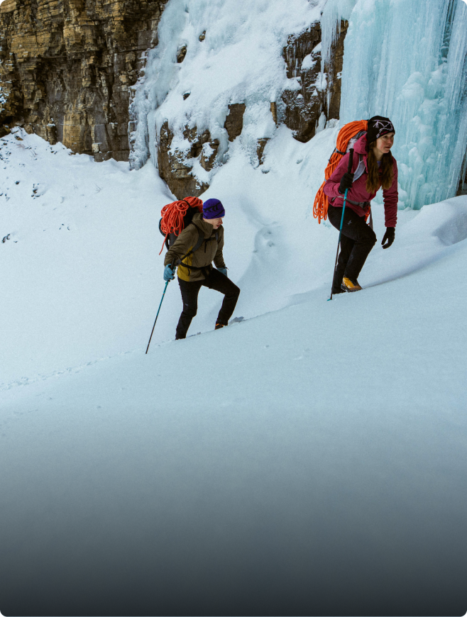 Two mountaineers with climbing packs and poles ascend a snowy slope near a frozen waterfall.