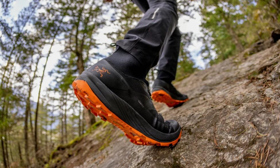 A person wearing black Arc'teryx trail shoes with bright orange soles walks up a rocky path in a forest.