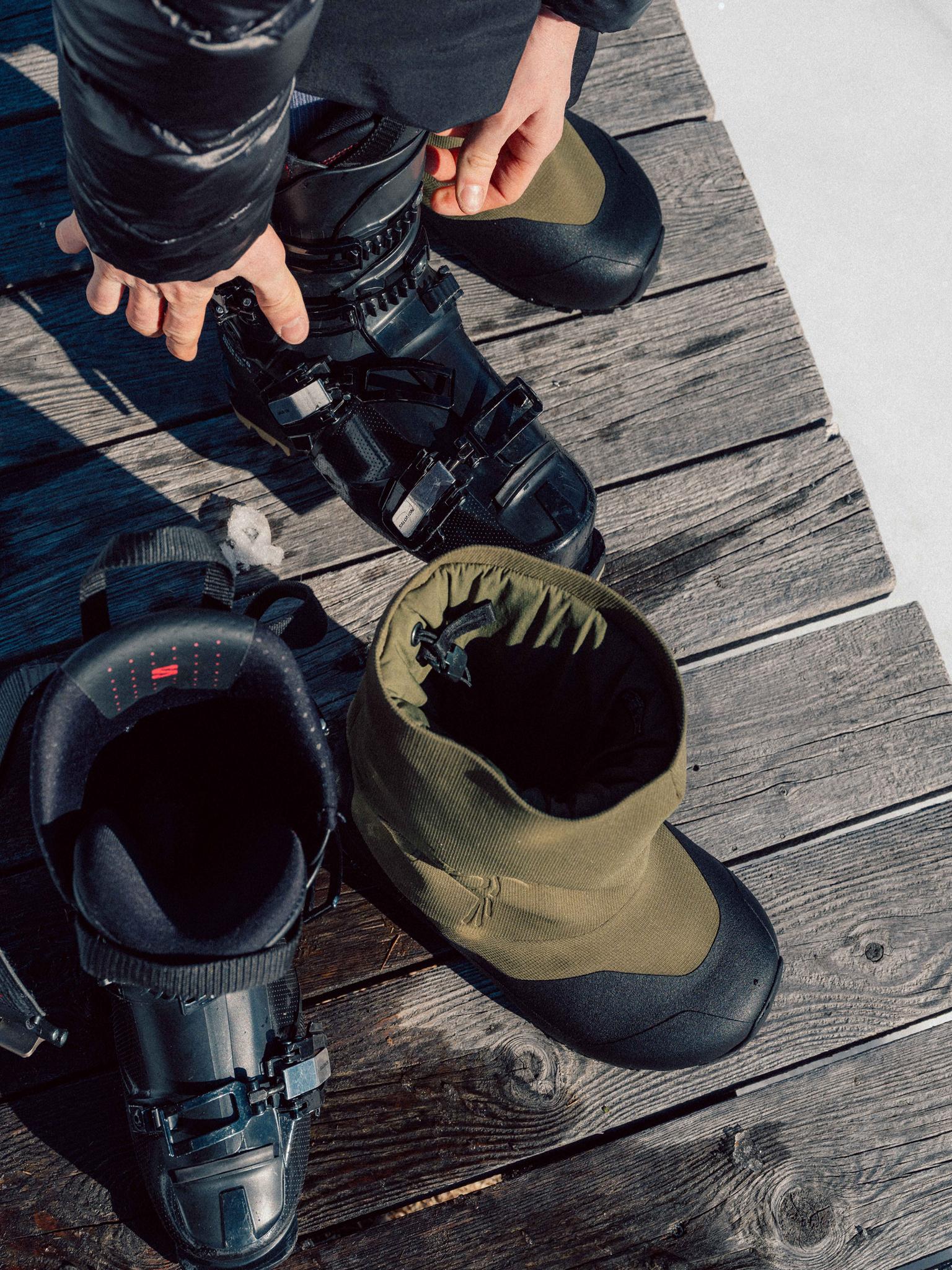 Hands adjusting a black ski boot on a wooden surface, next to an olive inner boot.