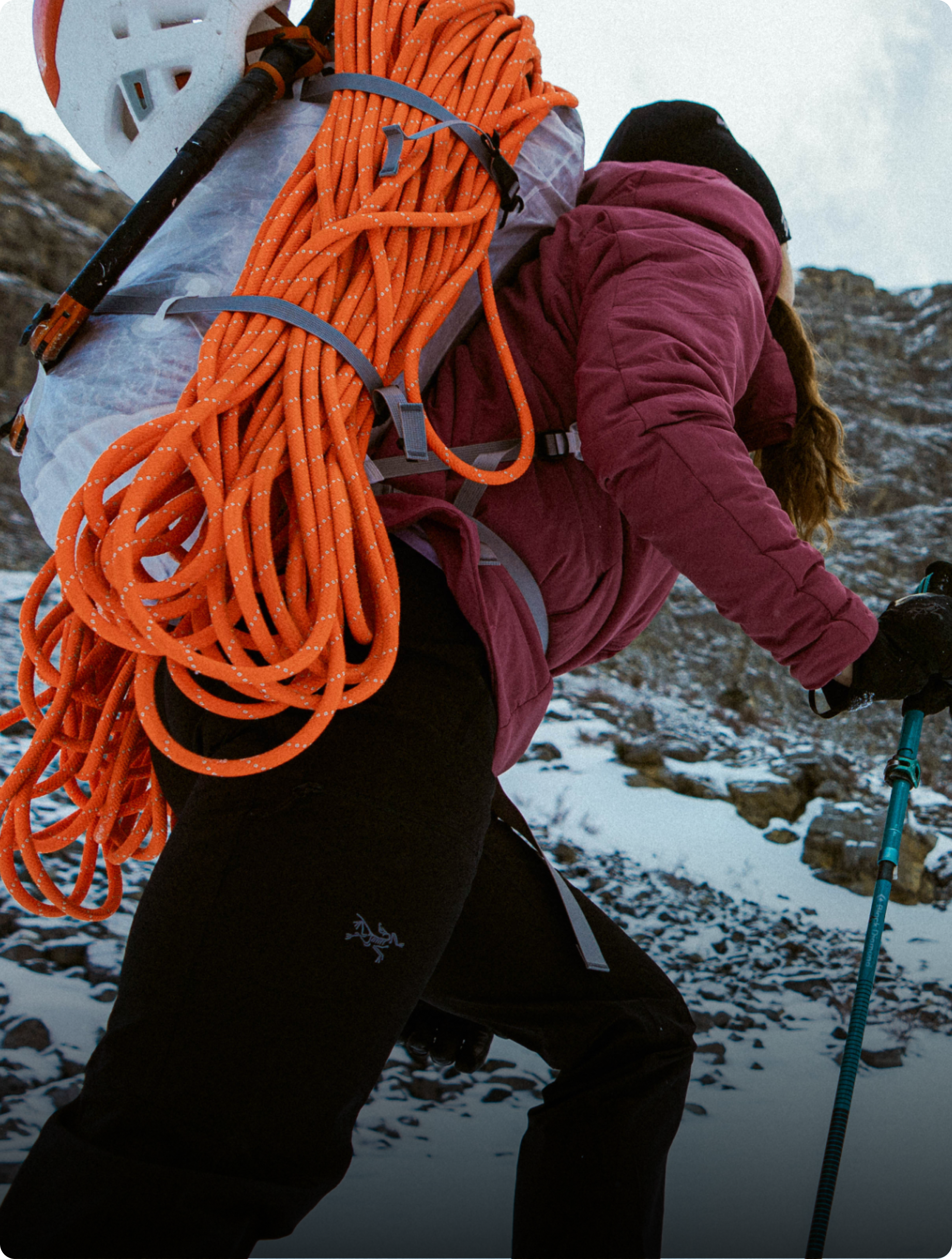 A person in winter climbing gear with an orange rope and ice axe on their backpack hikes up a snowy slope.