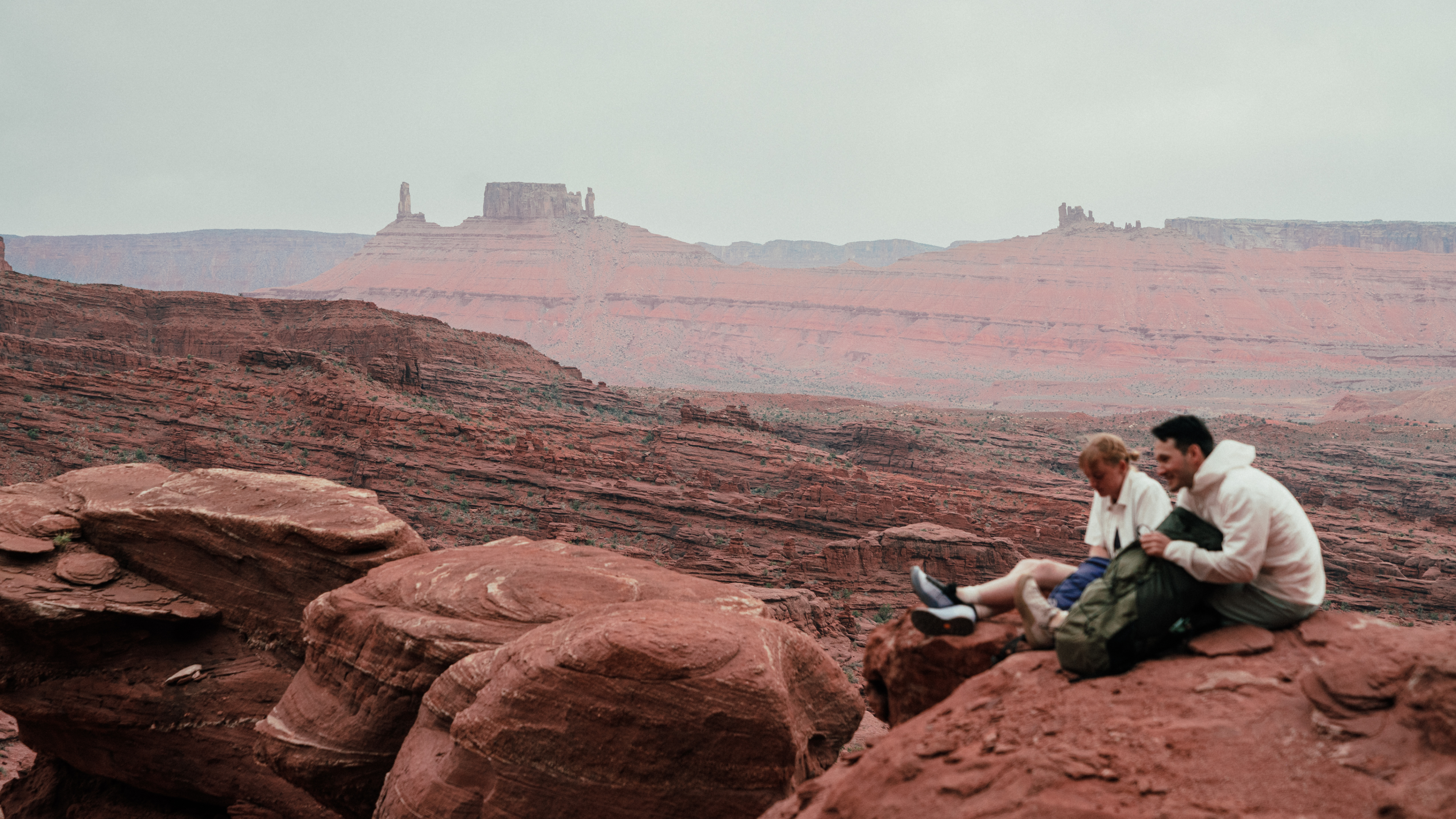 Two men sit on red rocks overlooking a vast red desert landscape with distant buttes under an overcast sky.