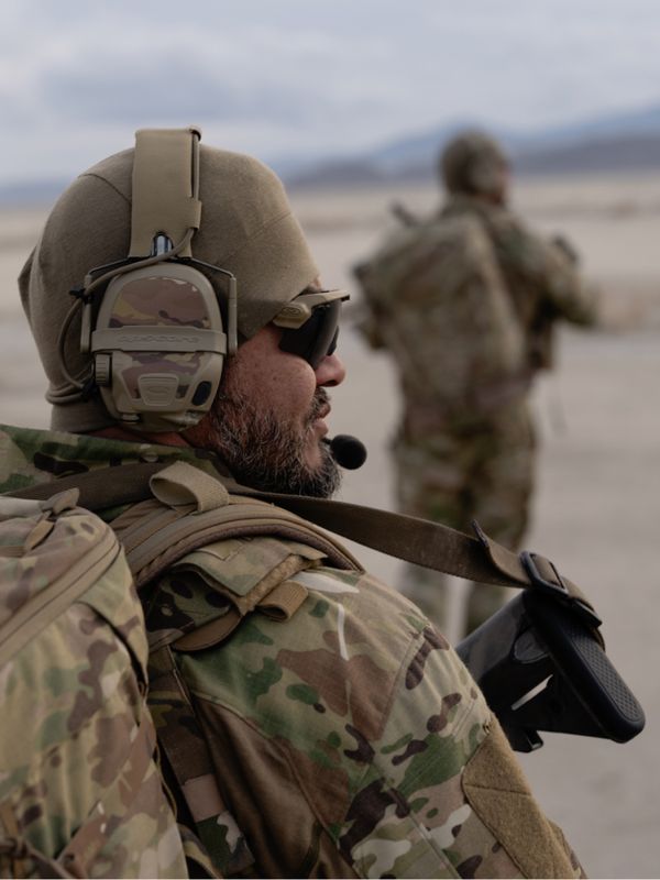 A bearded man in camouflage and tactical gear, including a headset and sunglasses, carries a rifle and backpack in a desert, with another person blurred in the background.