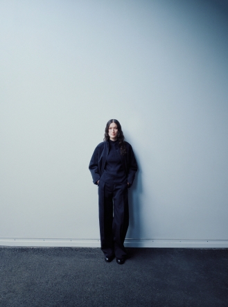 A woman with long dark hair wearing a dark jacket, turtleneck, and wide-leg pants stands against a plain light wall.